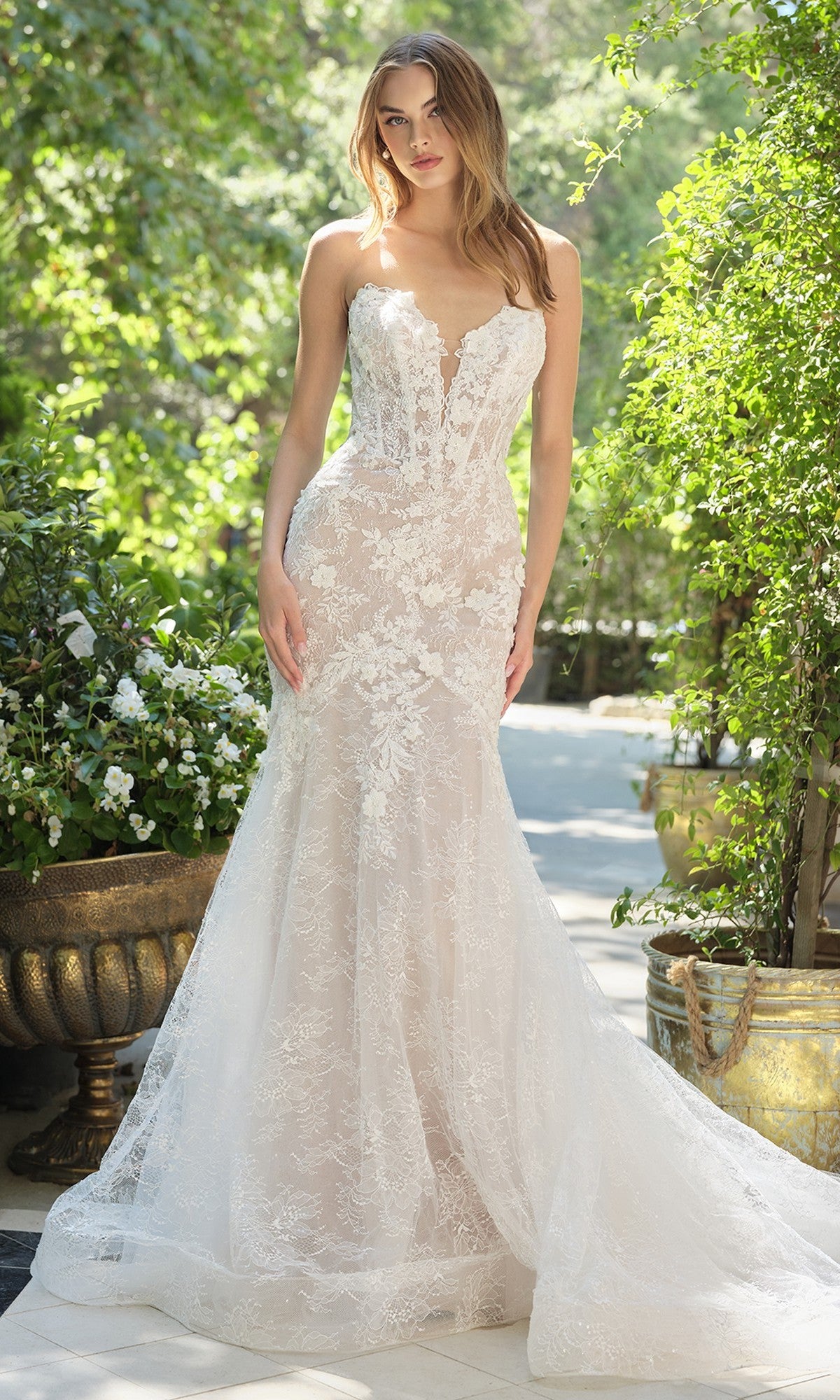 A woman stands outdoors among greenery wearing the Andrea and Leo WL072 long wedding dress, featuring a lace mermaid silhouette, boned corset bodice, and a long train.
